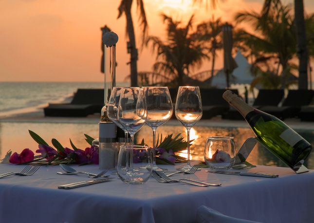 a dinner table, set for two, on the beach with the sun setting in the background.